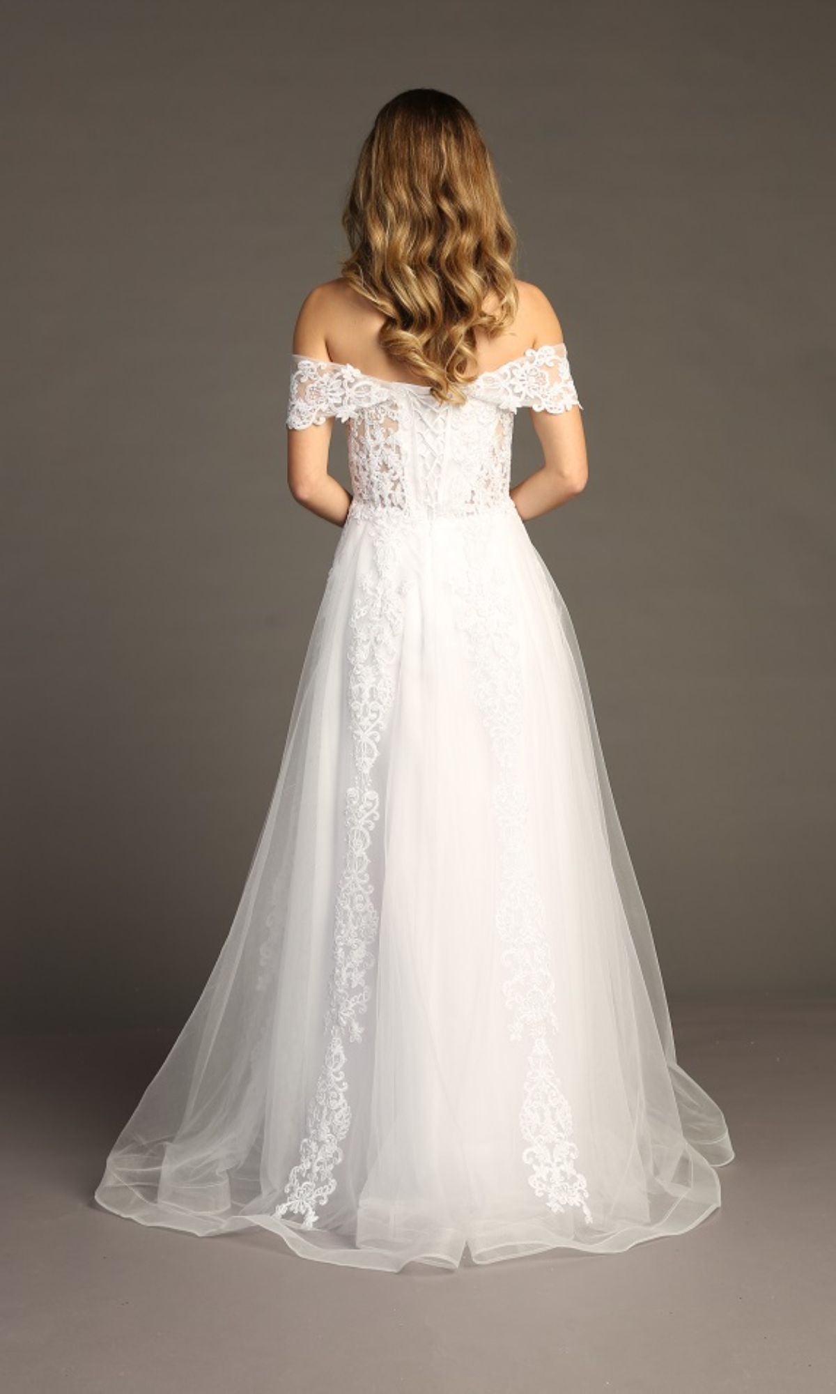 A woman with wavy hair faces away, wearing the Chicas CA1103 Long Lace Prom Ball Gown—an off-the-shoulder dress with a sheer, flowing train—posing against a plain background.